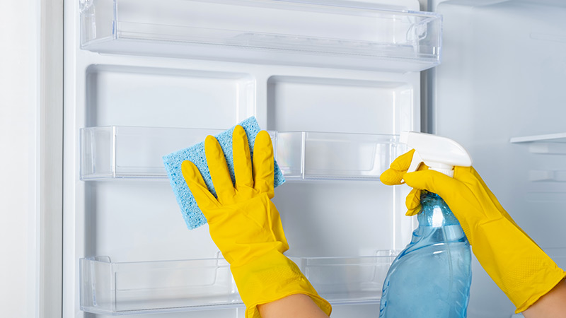 Cleaning refrigerator shelves with liquid spray and a blue sponge wearing yellow rubber safety gloves on hands.