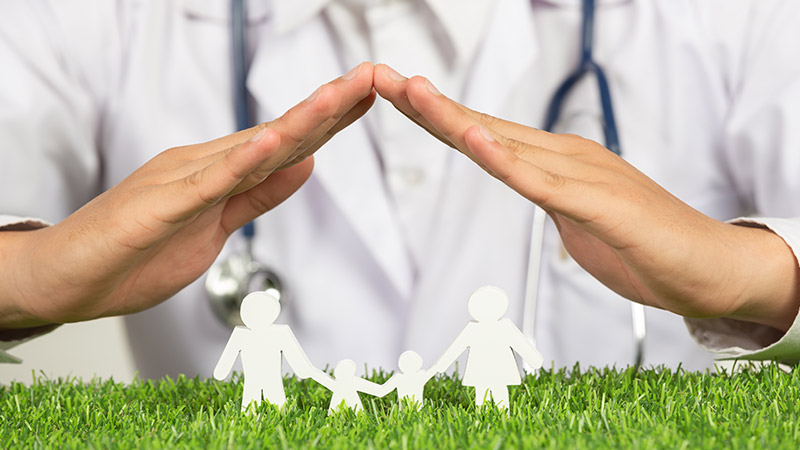Close-up picture of doctor's hands guarding a paper chain family of father, mother, son, and daughter on green grass.