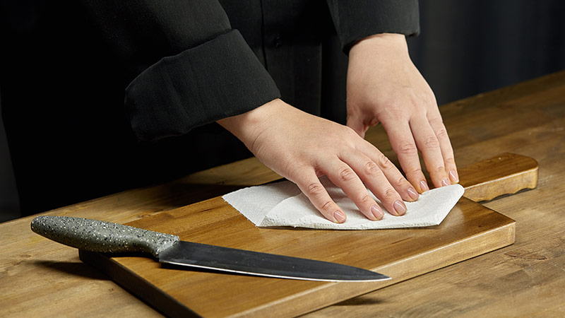 Hands wiping the cutting board with a paper towel while a knife placed on it. 