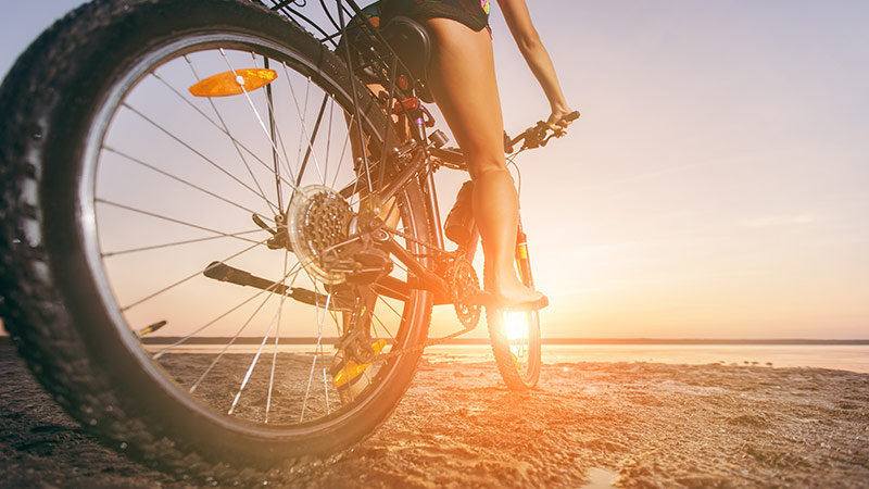 Woman in a multi-coloured outfit resting on her bicycle in a sand area near the lake.