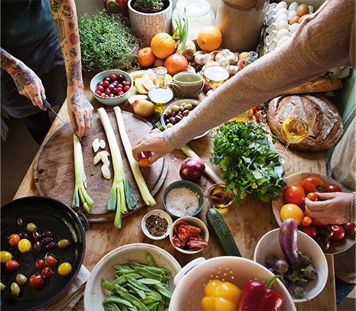 Two people taking food preparation on wooden table with varieties of food items. 