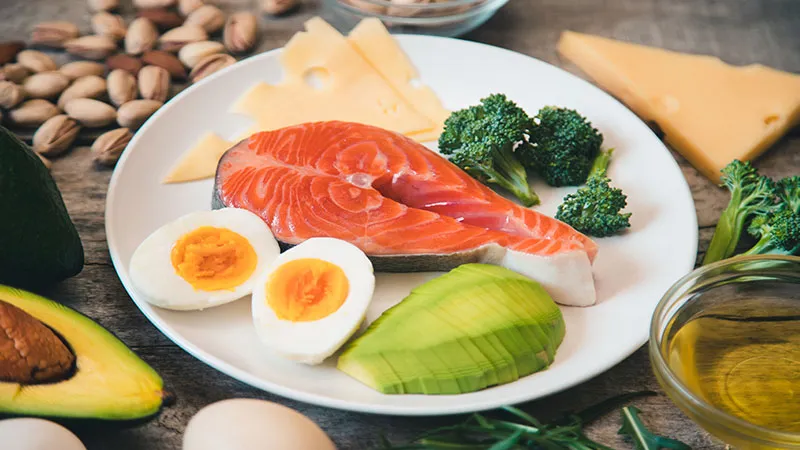 Food ingredients set with boiled egg, avocado, fish, broccoli, and cheese on wooden table.