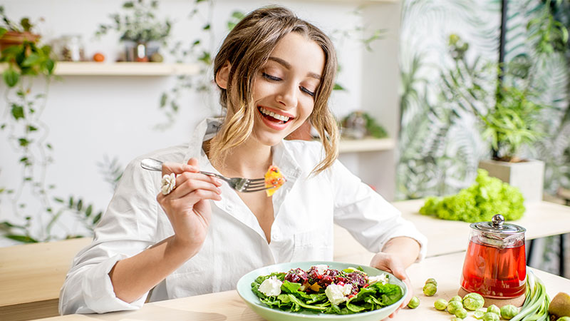 Young woman eating salad from a bowl while sitting in a beautiful interior with green flowers in the background.