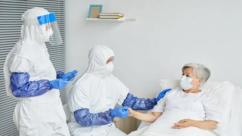 Two doctors in protective suits, respiratory masks, and gloves examine a senior woman in an infectious disease 