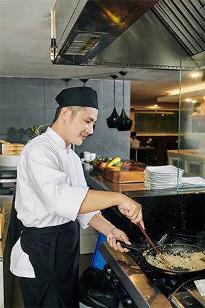 Young chef in uniform standing near the stove and frying rice on the pan in the kitchen