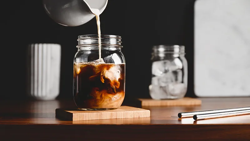 Pouring vegetarian soy milk into a glass jar with black coffee and ice cubes 