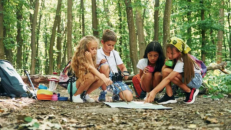 Kids conversing sitting on the ground in the forest, surrounded by travel equipment.