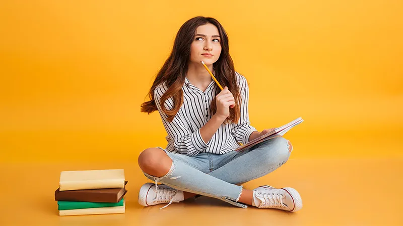 Young girl thinking while sitting on the floor studying with a bunch of books by her side