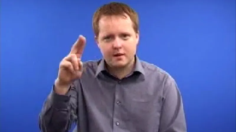 Sign language tutor seated in a room with his index and middle finger of the right hand attached and raised up