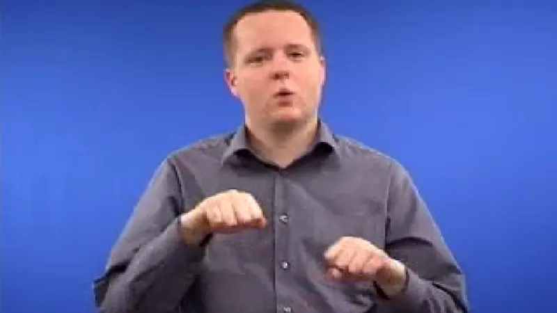 Sign language tutor seated in a room with blue background with both his hands curled up in a fist