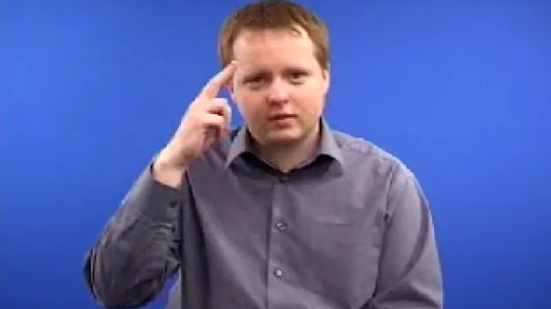 Sign language tutor seated in a room with his right hands raised near his head