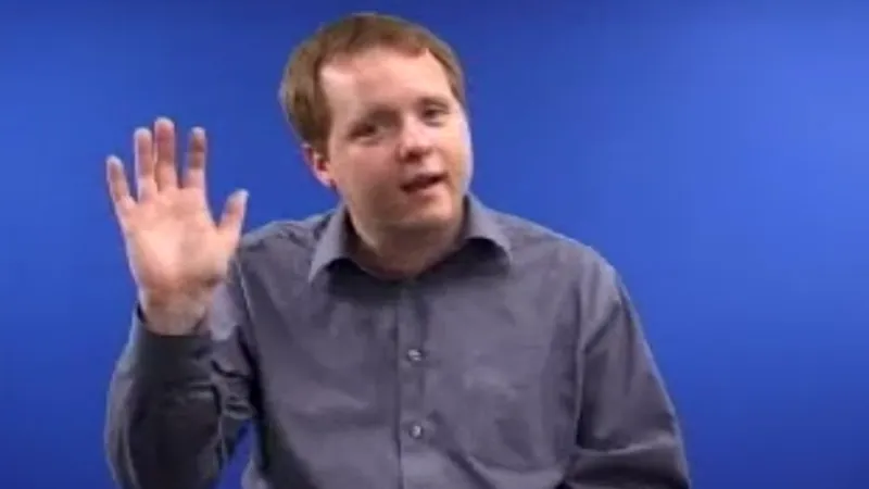 BSL tutor seated in a room with a blue background, with his right hand raised to sign Hi