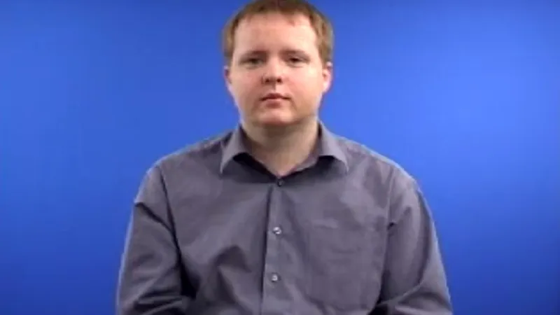 Male BSL tutor seated in a room with a blue background, with a normal posture