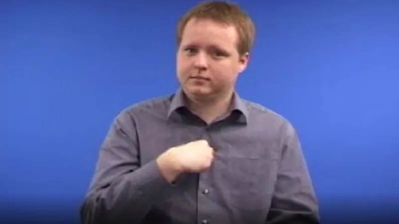 Male tutor seated in a room with a blue background, with his right hand curled up in a fist and placed near his chest