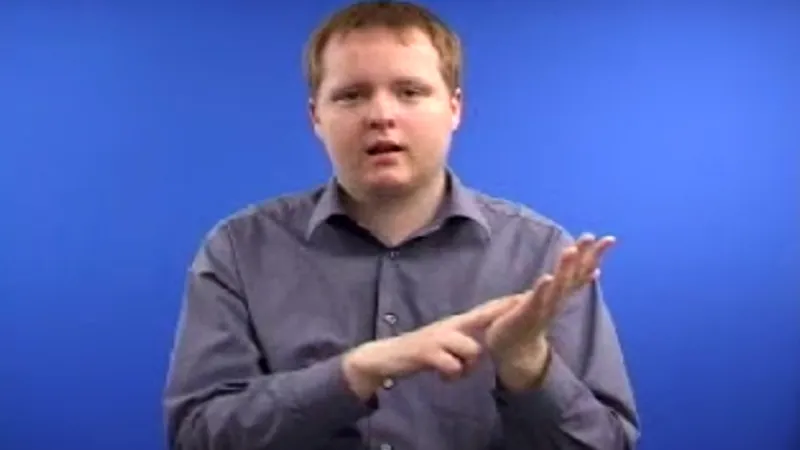 BSL tutor seated with his left hand raised palm facing upwards and index finger of right hand on the left palm