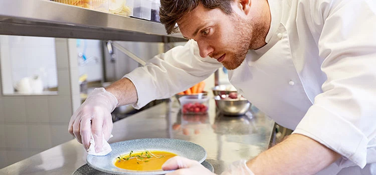 A male chef cook serving and cleaning plate of soup at restaurant kitchen