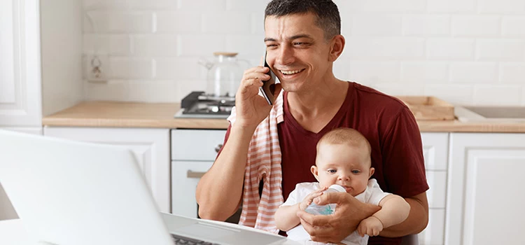 Father feeding his baby while having a pleasant conversation on the phone with a laptop in front of him.