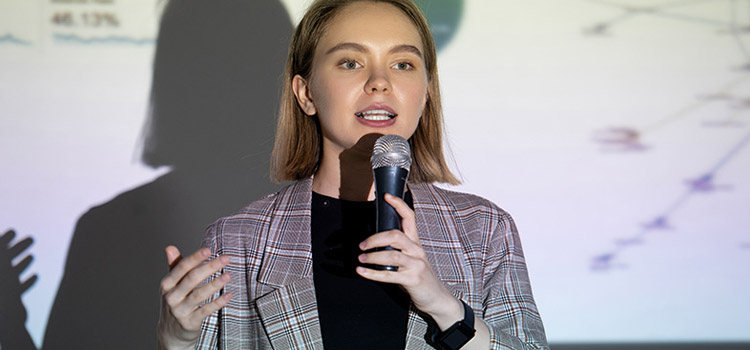 Confident woman delivering speech on a microphone along with a display on a background projector in the conference room.