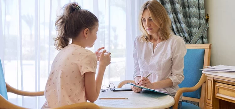 A female psychologist taking notes while her client- a little girl is sharing at a therapy session.