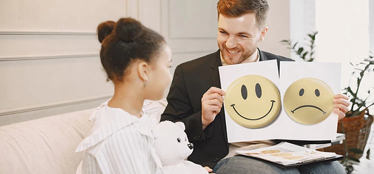 Little girl having a session with a psychotherapist who is showing her two pictures of happy and sad emoticons.