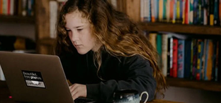 A girl working on her laptop, surrounded by a bookshelf and a cup of tea.