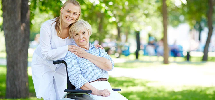 Female carer placing her hands on the shoulder of a happy elderly woman in a wheelchair while on outdoor wandering.
