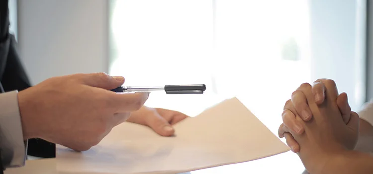 Suit-clad man giving a blank piece of paper and a pen to the person seated in front of him.