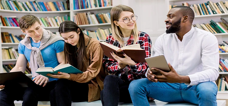 Four multiracial friends sitting in the library and studying using books and tablets. 