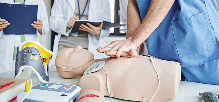 Partial view of paramedic practising chest compressions on a manikin close to a defibrillator.