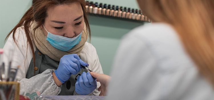 Nail stylist wearing mask and hand gloves decorating customer's fingernails 