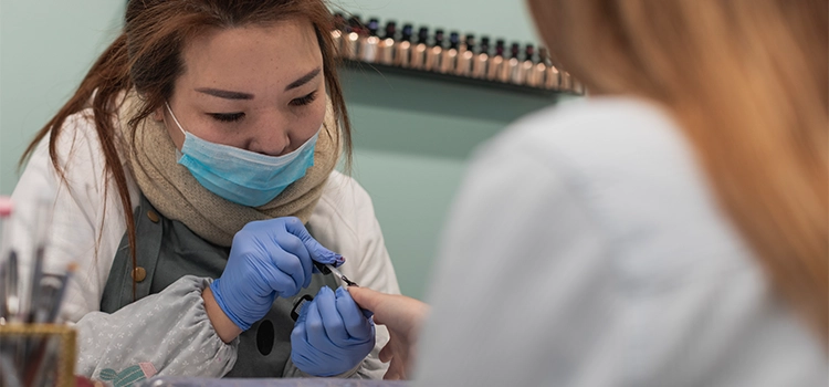 Nail stylist wearing mask and hand gloves decorating customer's fingernails