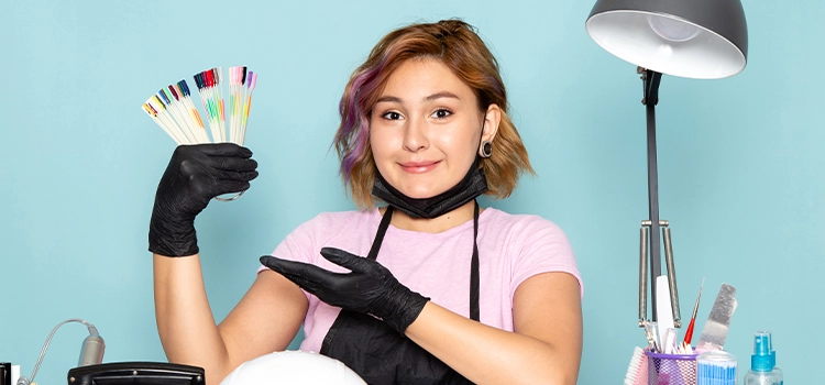 A nail technician wearing black gloves and a pink shirt showcases a set of nail colour samples while sitting at a manicure station.
